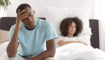 Hombre sentado al borde de la cama con cara de preocupación, en segundo plano una mujer con actitud de frustración.